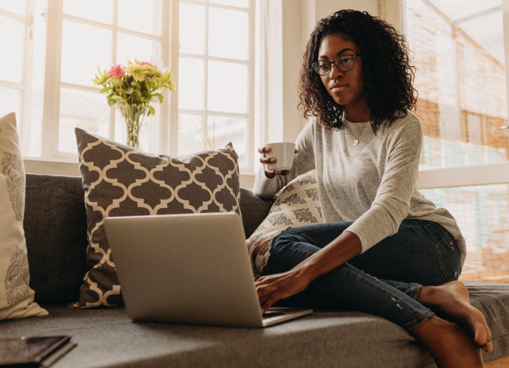 Woman using a laptop on her sofa and drinking coffee