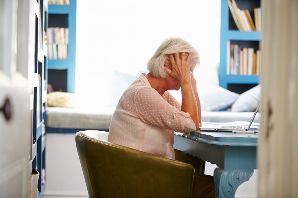 A stressed senior woman leans over her laptop computer and desk in a home office
