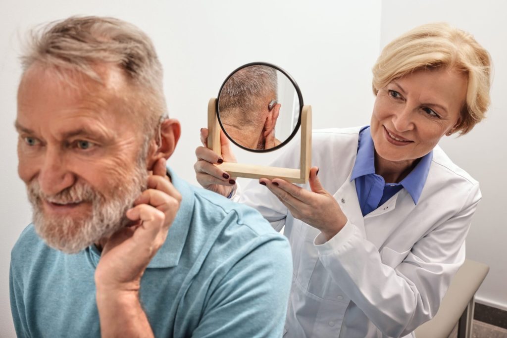 Elderly man trying on hearing aid in front of mirror with female audiologist putting hearing aid on ear during audiology consultation