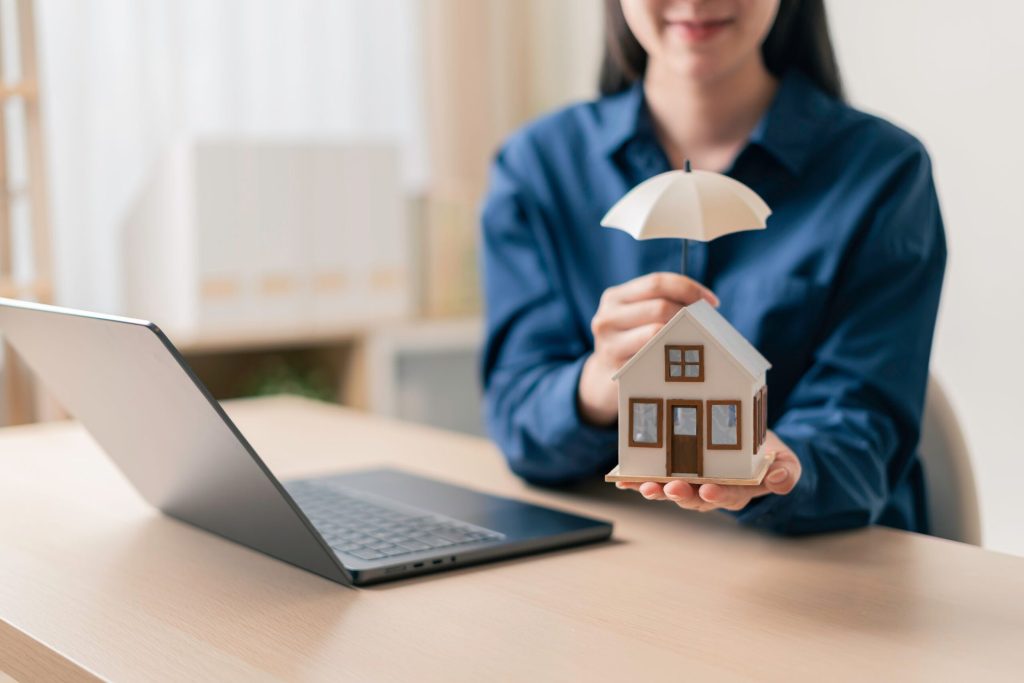 homeowner holding a miniature house under a small umbrella beside a laptop in a home office