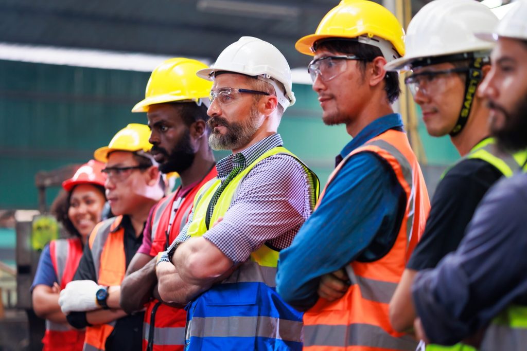 Team of workers in a factory or warehouse wearing safety vests, hard hats, safety glasses.
